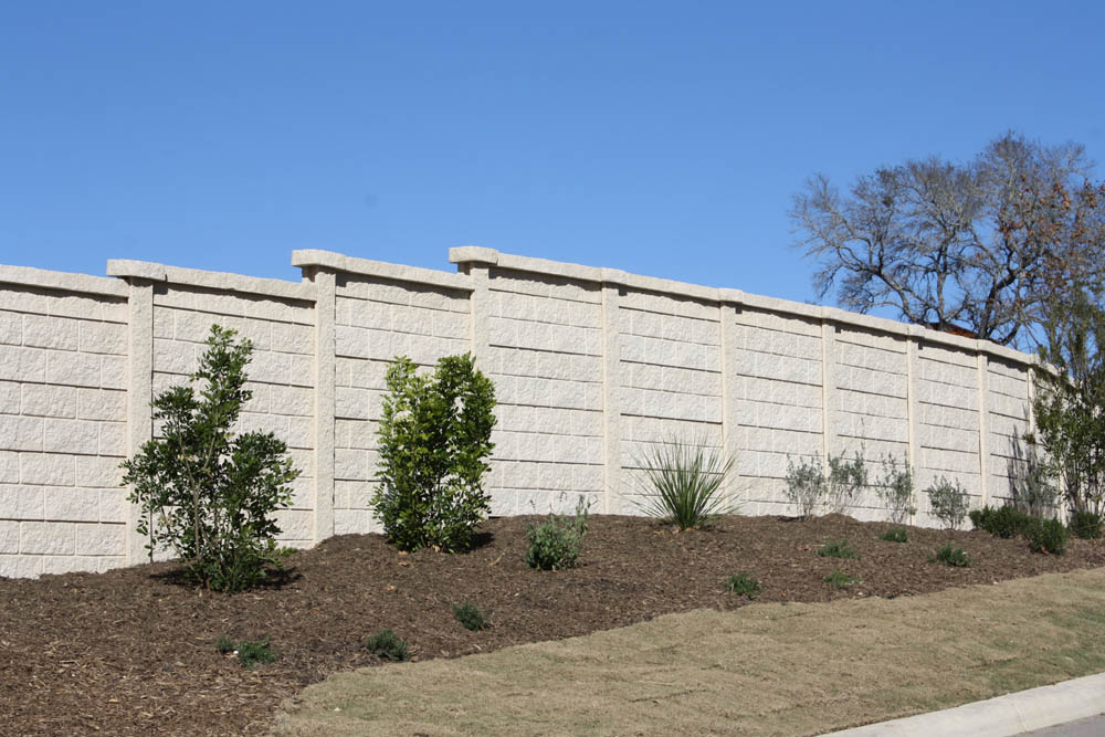 A tan concrete privacy wall runs along a sloped landscape with young shrubs and small plants in front of it, under a clear blue sky. A leafless tree is visible in the background on the right.