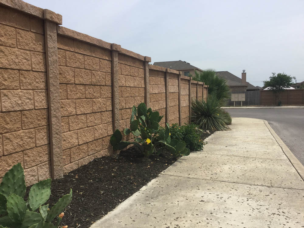 A sidewalk curves alongside a tan brick wall with prickly pear cactus and other desert plants growing in a mulched bed. Houses and a street are visible in the background under a cloudy sky.