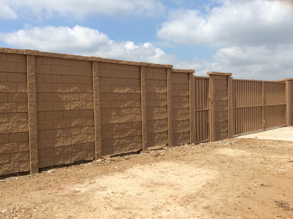 A tall brown concrete block wall with vertical support columns and a matching metal gate stands on a dirt ground under a partly cloudy sky.
