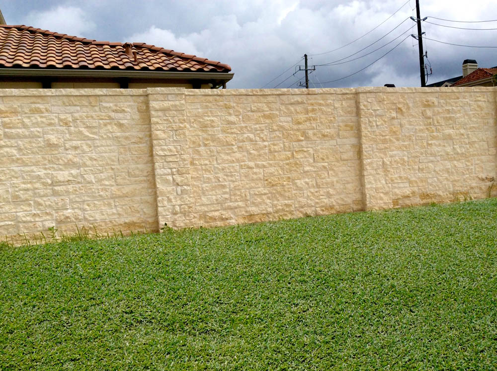 A beige stone wall runs across a grassy yard, with part of a house and its tiled roof visible on the left; power lines and a cloudy sky are in the background.