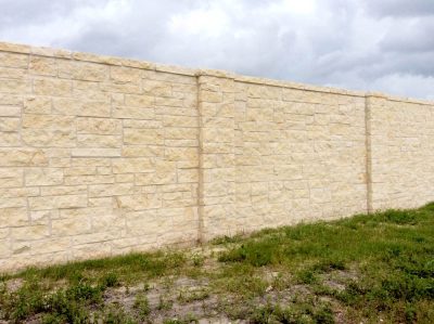 A tall, light-colored stone wall stands against a cloudy sky, with some green grass and patches of bare ground in front of it.