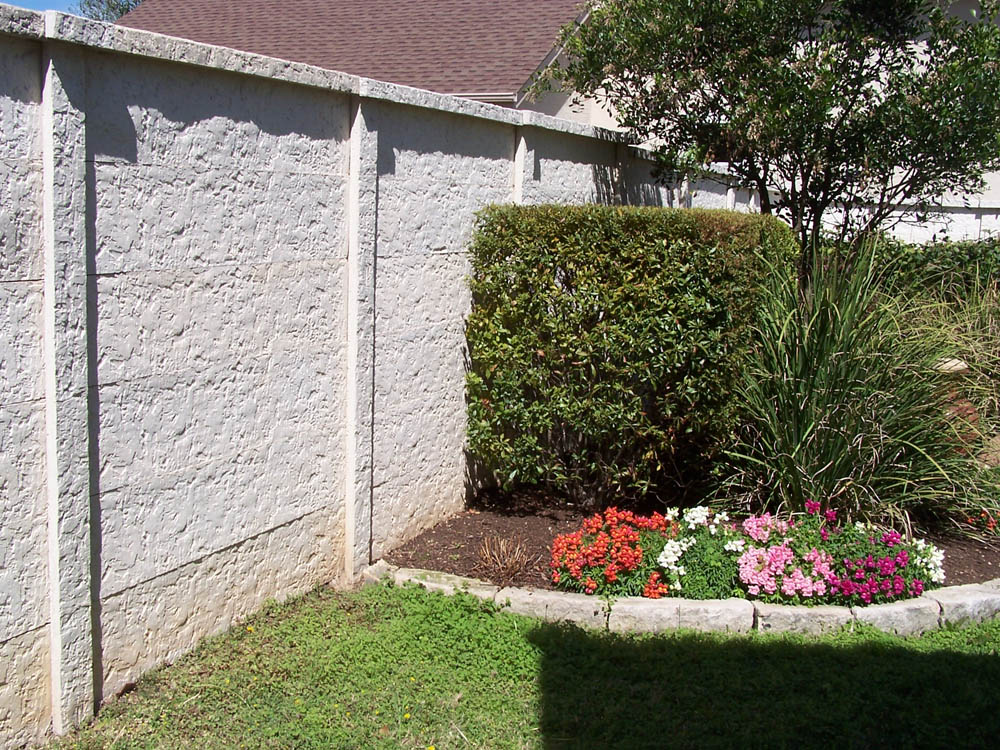 A neatly trimmed hedge, flowering plants, and ornamental grasses grow in a small garden bed bordered by stones next to a tall, white textured wall under bright sunlight.