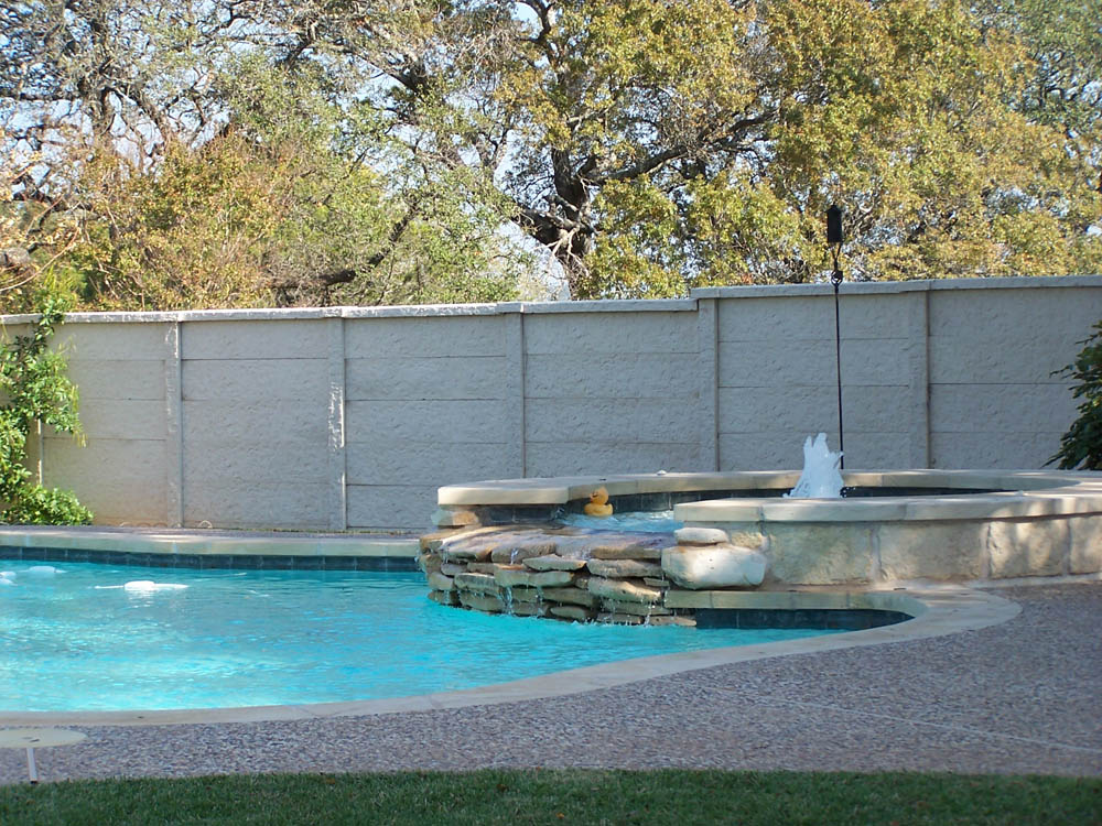 A backyard swimming pool with a small raised hot tub and waterfall feature. There’s a rubber duck in the hot tub, and trees and a tall stone fence in the background. The pool is surrounded by stone paving and grass.