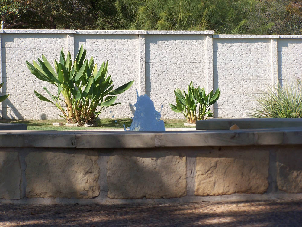 A small water fountain in a stone basin, with green plants and grass in the background, stands in front of a tall white brick wall under daylight.