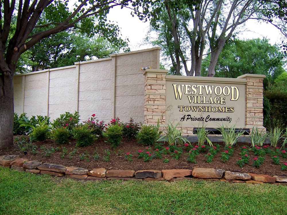 A landscaped entrance sign for Westwood Village Townhomes, A Private Community, with flowers, shrubs, and trees in front of a stone wall.