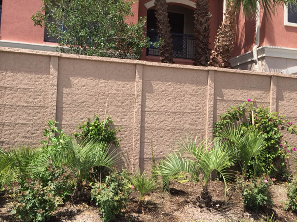 A beige concrete wall with green shrubs and small palm plants in front, set against a building with a reddish exterior and tall windows partially visible behind the wall.