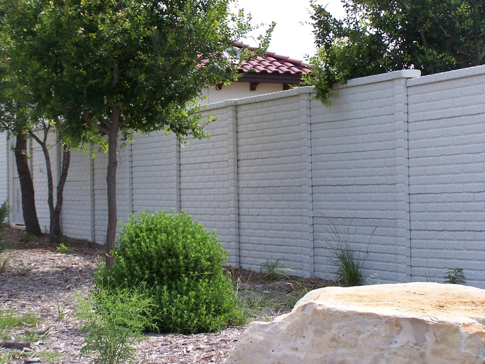 A tall white brick wall runs alongside trees, bushes, and a large pale rock, with part of a red-tiled roof of a house visible above the wall in the background.