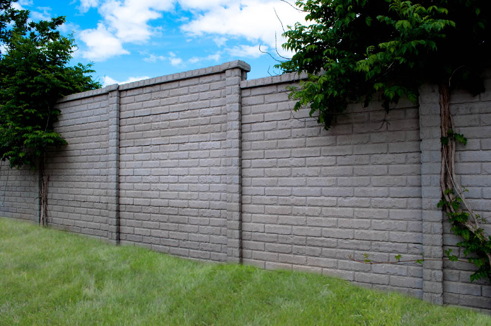 A tall, light gray brick wall runs across a grassy area, with green leafy vines climbing and hanging over the top in several spots. Blue sky with white clouds is visible above.