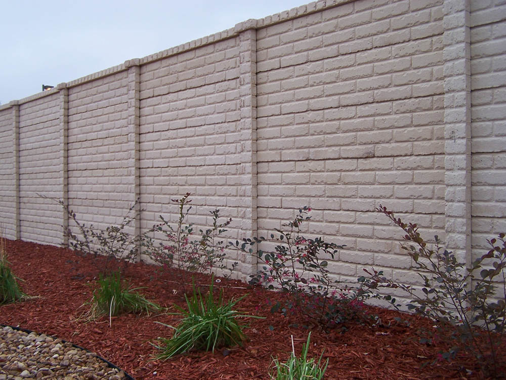 A beige brick wall with vertical columns stands behind a mulched garden bed, which contains small shrubs, grasses, and decorative rocks. The sky above is overcast.