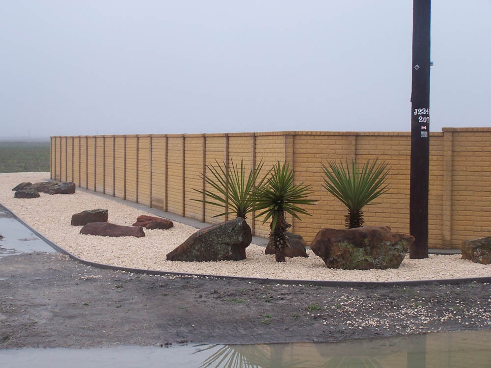 A landscaped area with tan gravel, large rocks, and several small palm-like plants sits beside a tall yellow brick wall on a foggy day. A utility pole stands in the gravel near the plants.