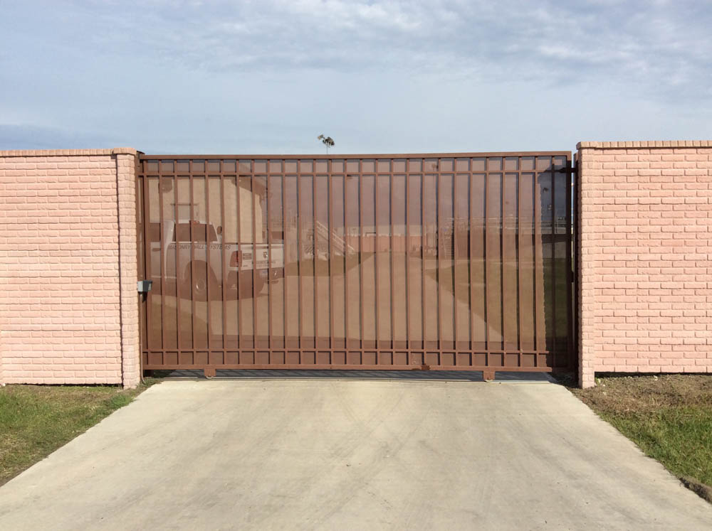 A brown metal gate with vertical bars is set between two light pink brick walls, blocking the entrance to a paved driveway. A white vehicle is visible through the bars and behind the gate. The sky is partly cloudy.