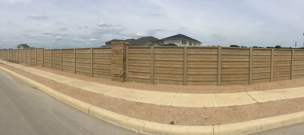A long, beige wooden fence with stone pillars runs alongside a sidewalk and street, enclosing the backyard of a suburban house. The sky is mostly cloudy.