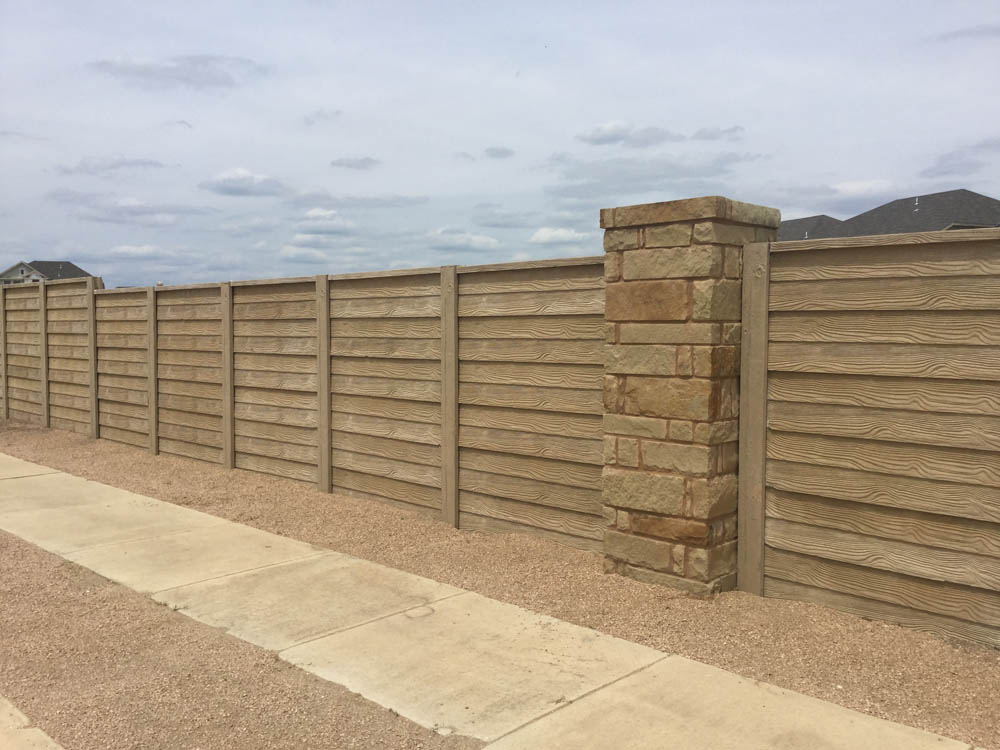 A long wooden privacy fence with horizontal panels and a stone pillar runs alongside a gravel path and a concrete sidewalk under a cloudy sky.