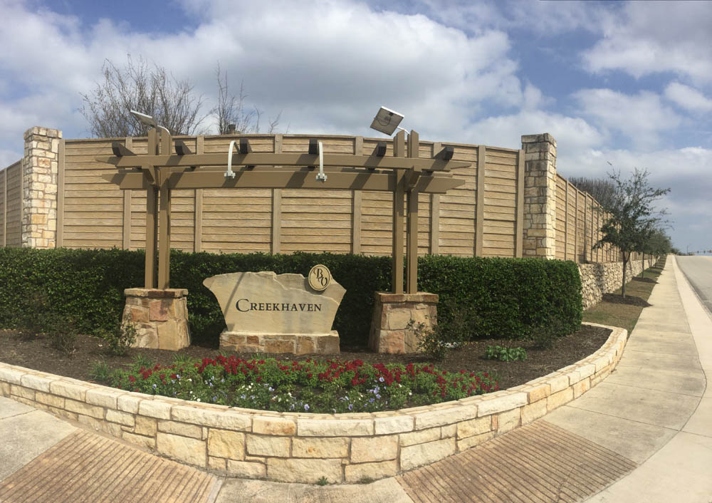 A stone sign reading “Creekhaven” stands in front of a wooden fence, surrounded by bushes, flowers, and a pergola-like structure at the entrance to a residential neighborhood.