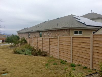 A wooden privacy fence runs along the backyard of a brick and beige house with a sloped roof under a cloudy sky. Some shrubs and dry grass are visible in the yard.