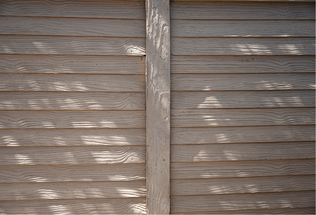 Sunlight casts dappled shadows of leaves onto a beige, wooden plank fence with horizontal panels and a vertical support beam in the center. The wood grain and texture are clearly visible.