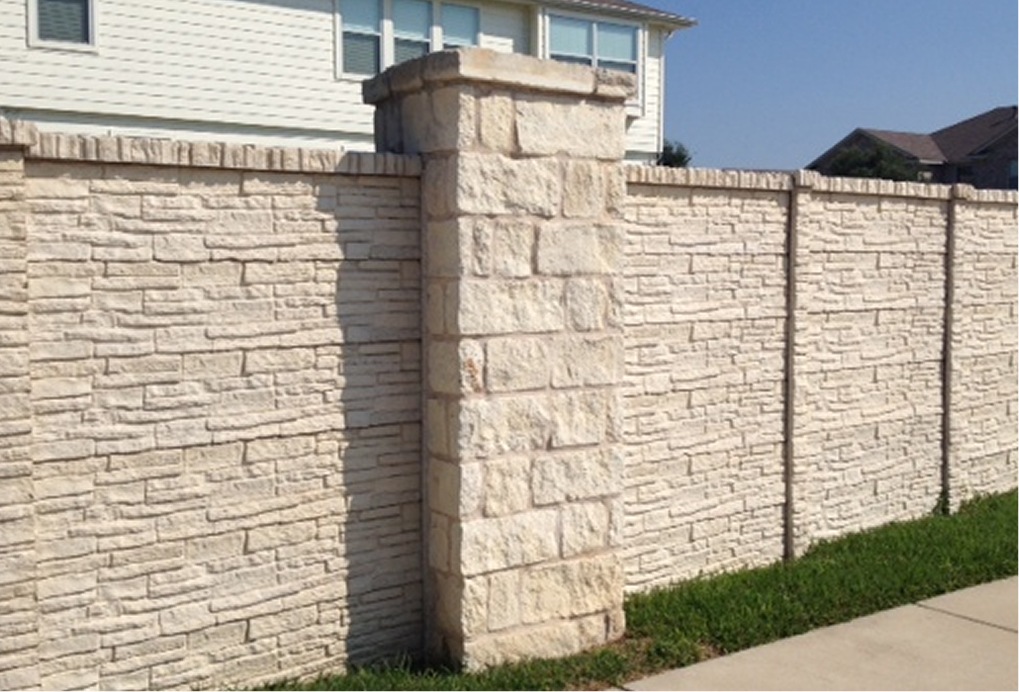A light-colored stone wall with a large, square pillar stands alongside a sidewalk, with green grass at the base and parts of houses visible in the background under a clear blue sky.