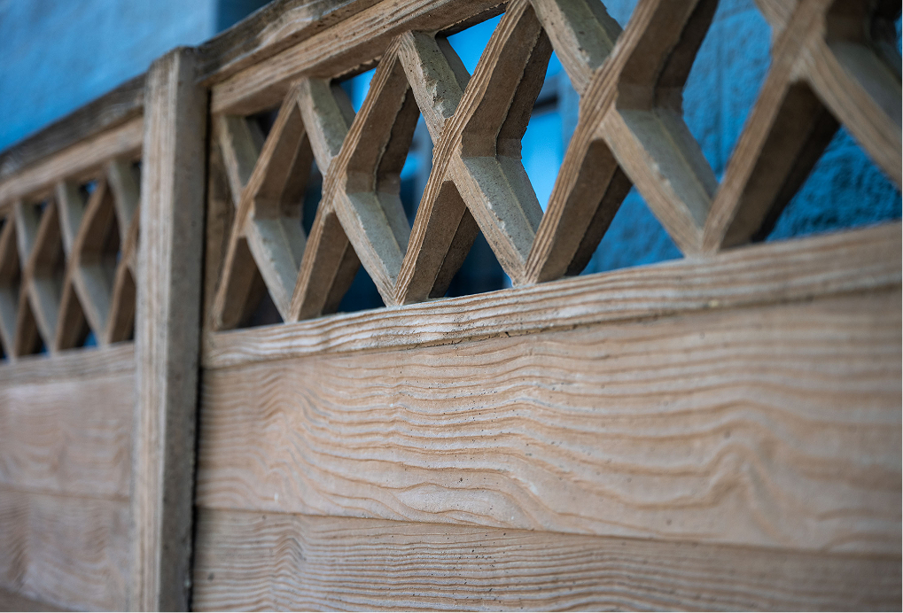 Close-up of a wooden fence with a lattice pattern of diamond-shaped openings at the top, and horizontal wooden planks with visible grain patterns below. Blue background visible through the lattice.