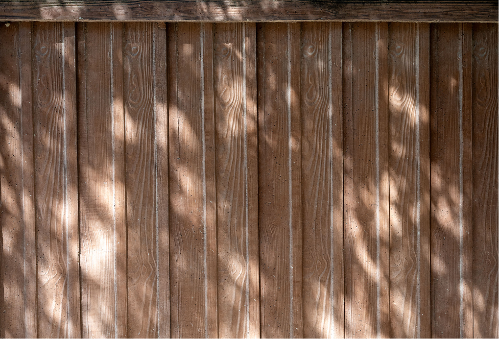 Vertical wooden planks with a natural brown finish; sunlight filters through leaves, casting irregular, dappled shadows across the surface.