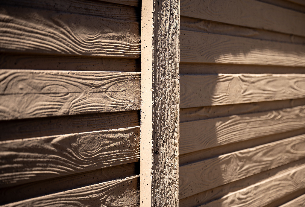 Close-up of tan-colored, textured wooden siding panels with visible wood grain patterns and shadows, divided vertically by a rough wooden post in the center.