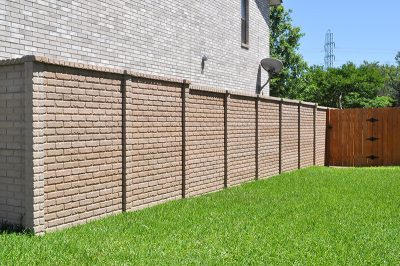 A beige brick privacy wall runs alongside a house, separating a green lawn from a wooden gate. Trees and a power line tower are visible in the background under a clear blue sky.