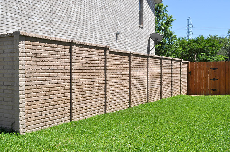 A beige brick privacy wall runs alongside a house, separating a green lawn from a wooden gate. Trees and a power line tower are visible in the background under a clear blue sky.