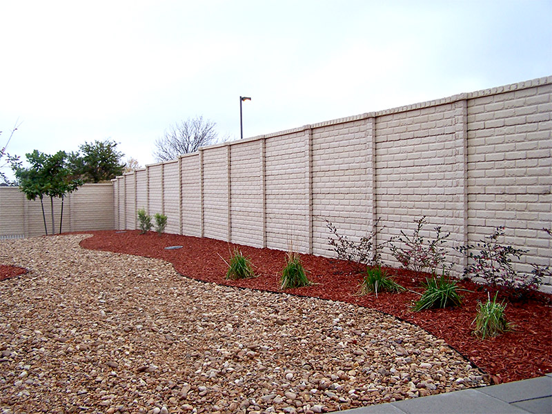 A yard with a tall beige brick wall, decorative gravel and mulch, and small green plants arranged along the mulch near the wall, under a cloudy sky.