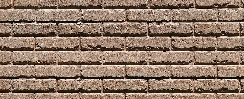 A close-up view of a beige brick wall, showing horizontal rows of rectangular bricks with rough textures and visible mortar lines between them.