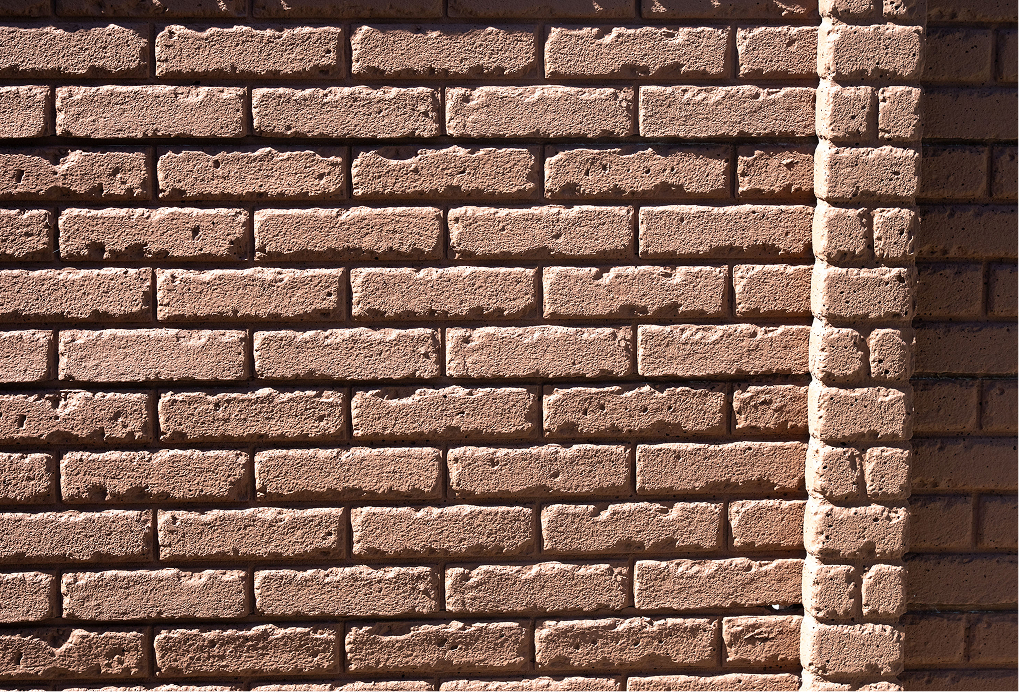 Close-up of a brown brick wall with sunlight casting shadows. The bricks are arranged in horizontal rows, and a vertical corner is visible on the right side of the image, emphasizing the wall’s texture and structure.