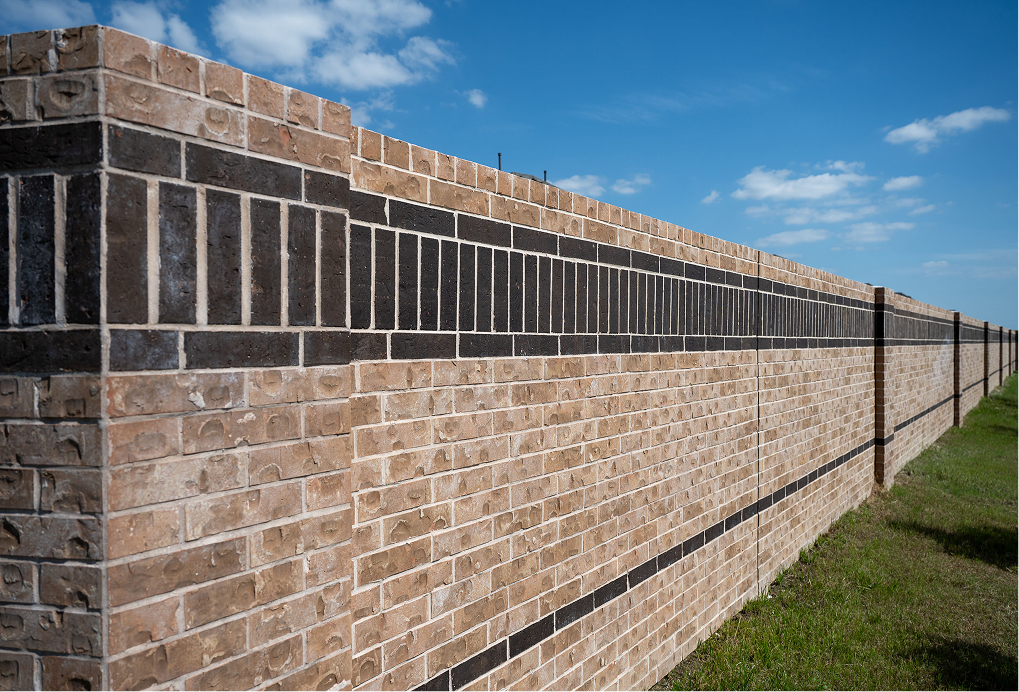 A long brick wall with a horizontal dark stripe runs through a grassy area under a bright blue sky with scattered white clouds.