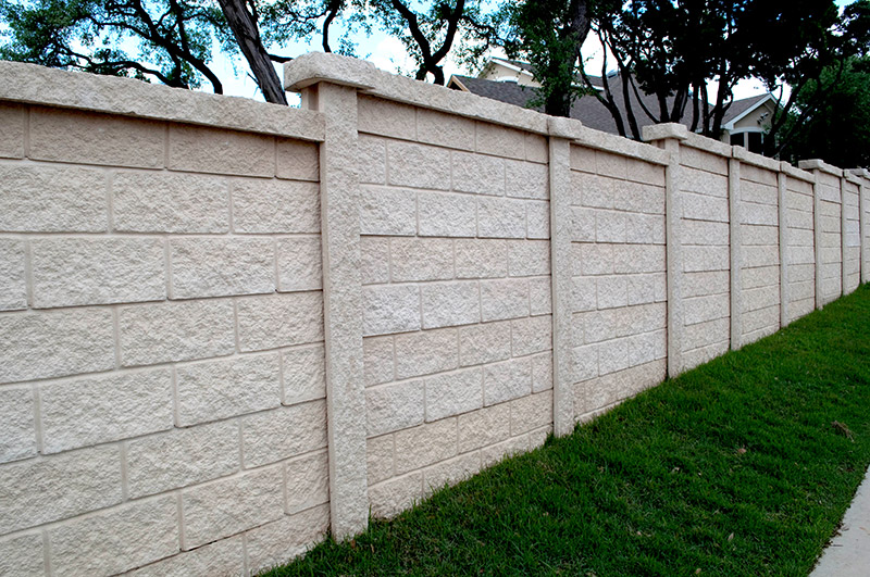 A tall beige concrete block fence runs along a well-maintained grassy area, with trees and houses visible behind the fence in the background.