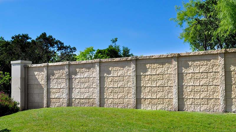 A beige stone masonry wall with textured rectangular blocks stands on a grassy lawn, backed by trees and greenery under a clear blue sky.
