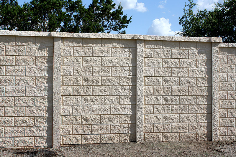 A tall, beige concrete block wall with a textured brick pattern stands outdoors in front of some green trees under a blue sky with scattered clouds.
