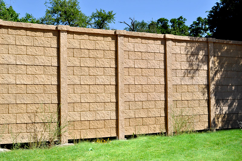 A tall beige concrete block wall with vertical supports stands behind a patch of green grass and sparse weeds, with trees and a clear blue sky in the background.