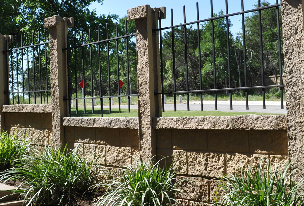 A stone and metal fence with vertical black bars stands in front of a road lined with trees and greenery. Tall decorative grass grows along the base of the fence in the foreground.