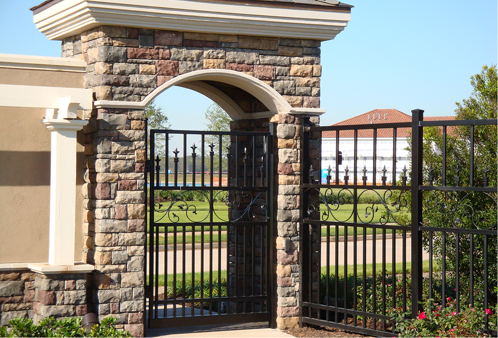 A decorative wrought iron gate set in a stone archway opens to a manicured lawn, with a large building and clear blue sky visible in the background.