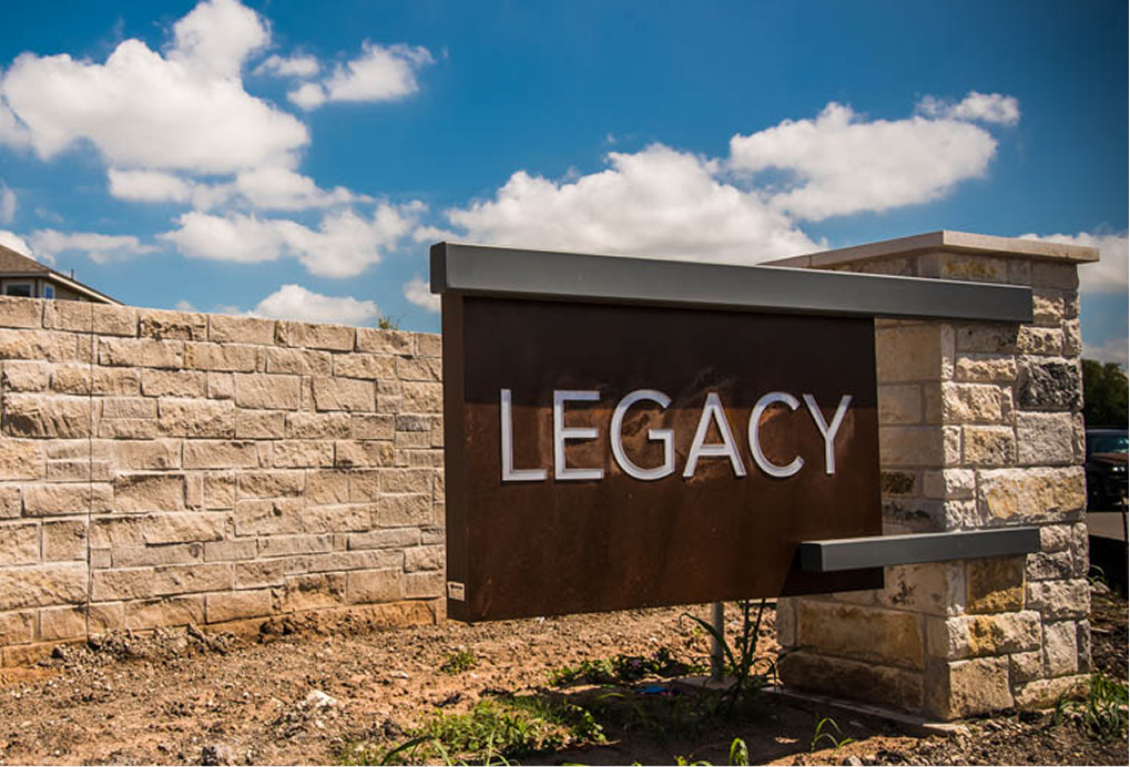 A large metal sign reading LEGACY is mounted on stone pillars next to a stone wall, under a bright blue sky with scattered clouds.