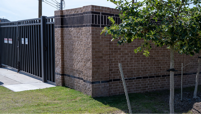 A brick wall with a black metal gate runs along a sidewalk. A small tree grows in the grassy area next to the wall, and signs are posted on the gate. Sunlight casts clear shadows on the wall and grass.