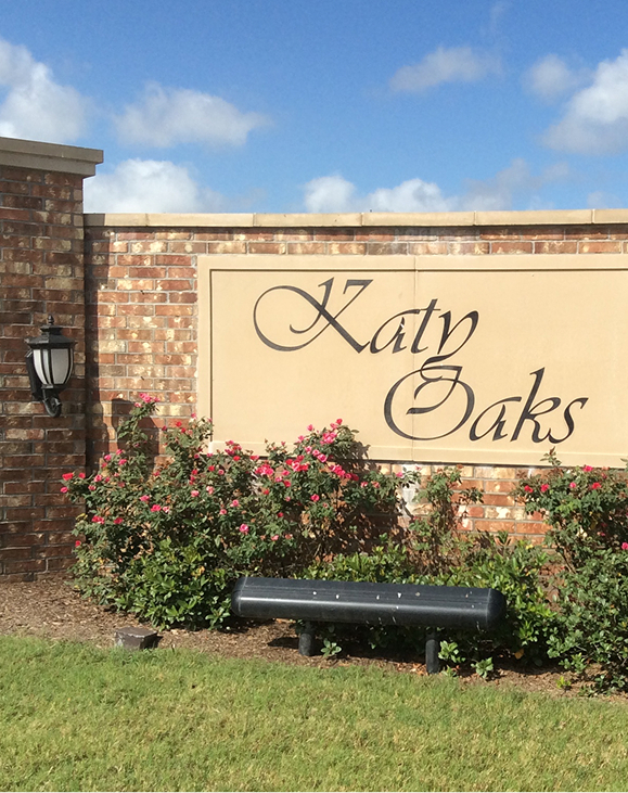 A brick wall sign with black script reading Katy Oaks is surrounded by pink flowering bushes and a black lamp, with grass and blue sky in the background.