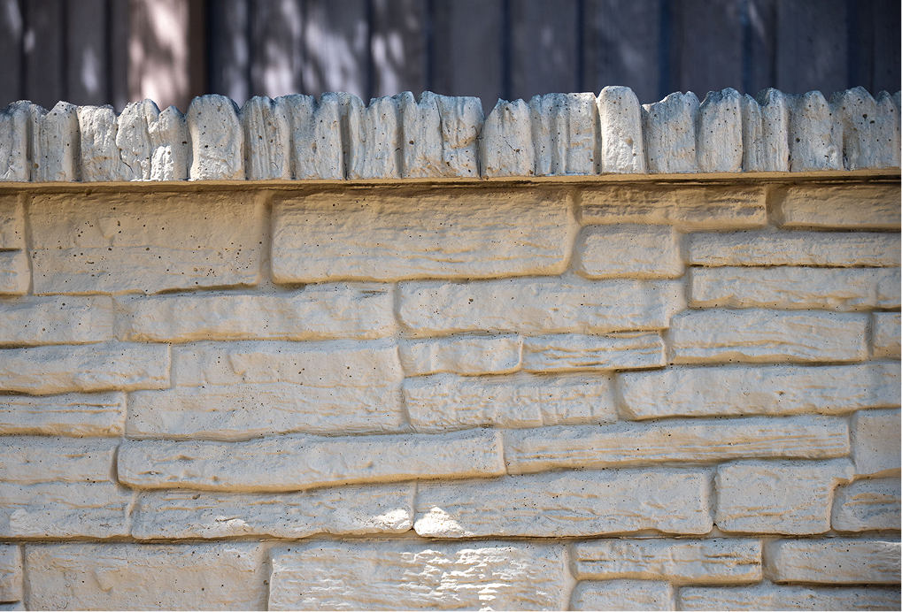 Close-up of a beige stone wall with a textured, irregular stone pattern and a rough, vertical stone edge on top. Soft shadows from tree branches are visible on the upper part of the wall.