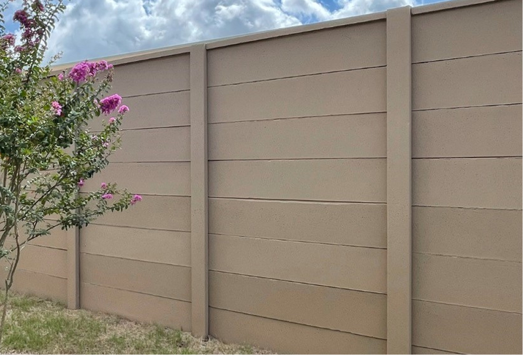 A tall beige concrete fence stands in front of a partly cloudy sky, with a small tree featuring pink flowers on the left side of the image.
