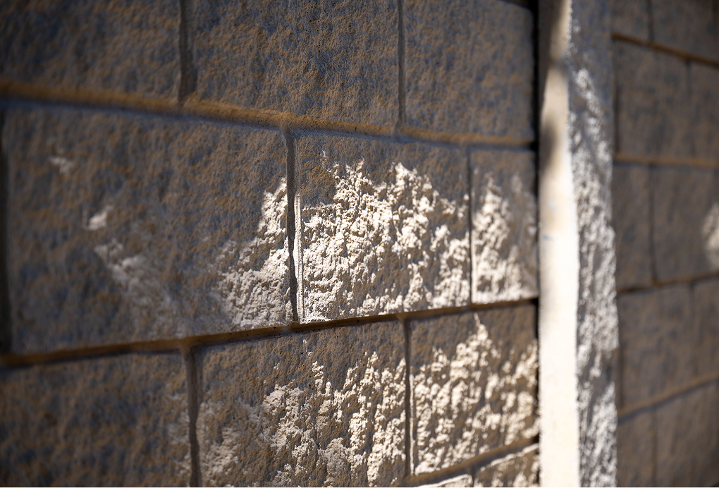 Close-up view of a sunlit textured stone or concrete block wall, with light and shadow creating contrasting patterns across the rough surface. A vertical line separates two sections of the wall.