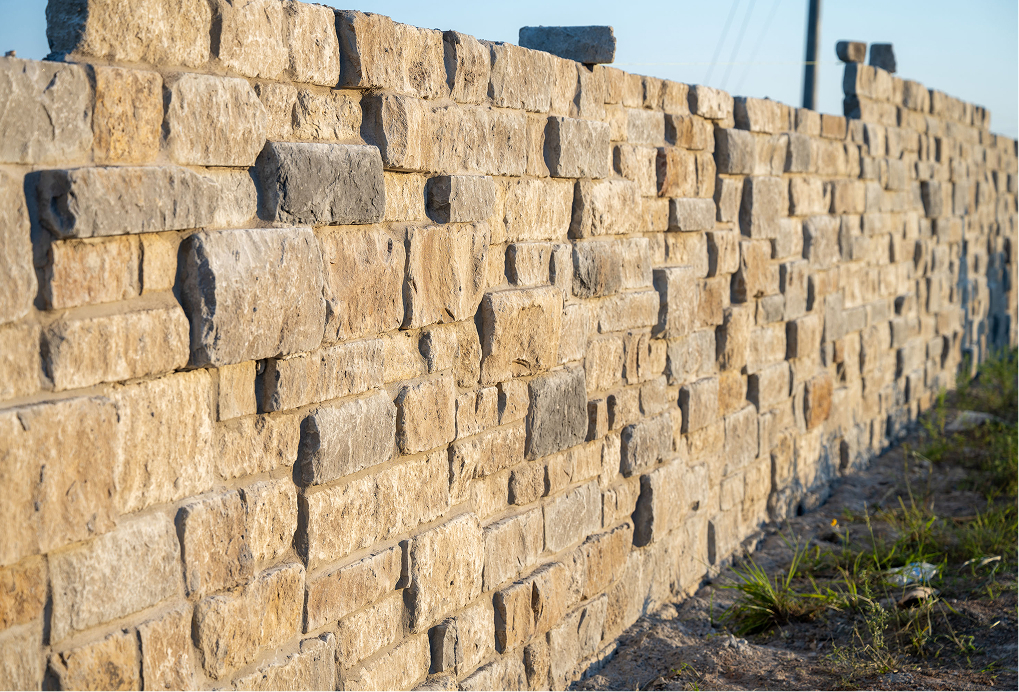 A close-up view of a long, uneven stone wall made of rectangular tan and gray bricks, with some grass and dirt visible at the base, stretching into the distance under a clear sky.
