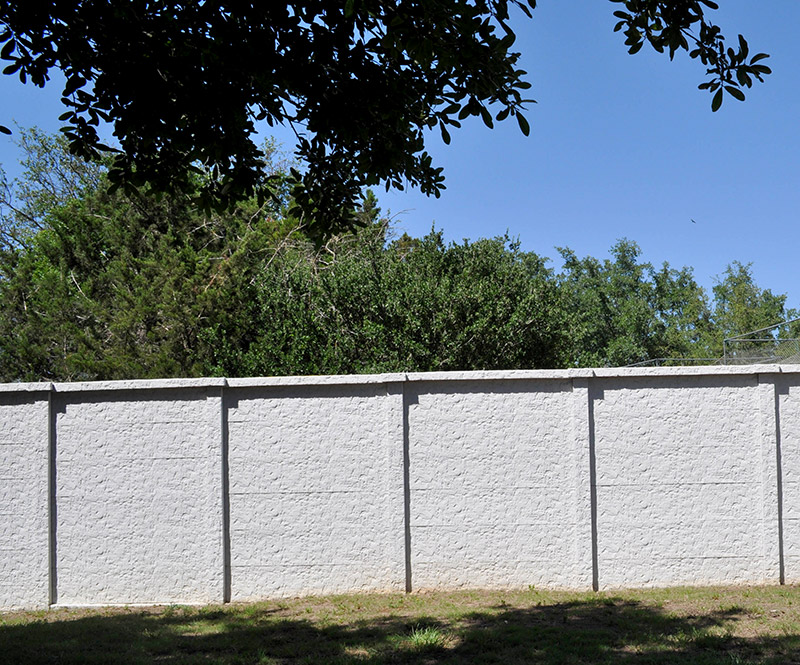 A tall, light gray concrete wall stands in front of dense green trees under a clear blue sky, with tree branches and shadows visible in the foreground.