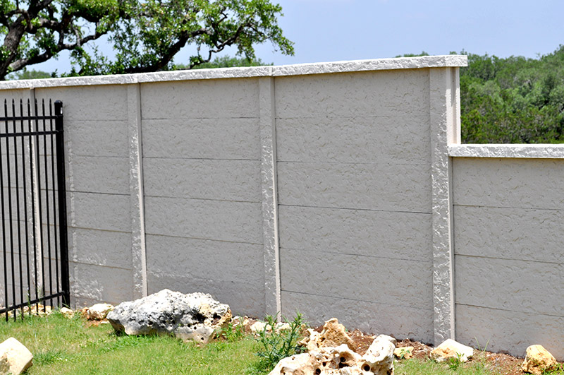 A beige textured concrete wall with vertical supports stands next to a black metal fence; rocks and green grass are in the foreground, and trees are visible in the background.