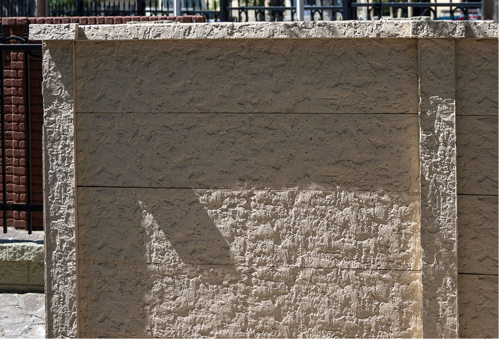 A textured beige concrete wall stands in sunlight, casting angular shadows. Behind it, black metal railings and red brick structures are partially visible at the top of the image.