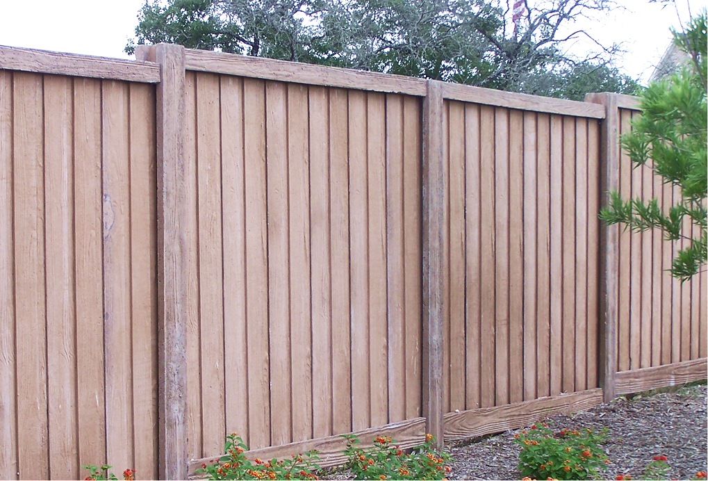 A tall wooden privacy fence with vertical panels and sturdy posts stands in a yard, with green bushes and small orange flowers in the foreground and leafy trees in the background.