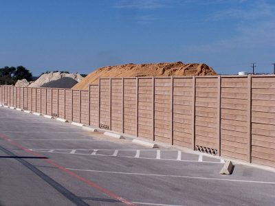 A tall, light brown wooden fence runs alongside a paved parking lot with painted parking spaces. Sand or gravel piles are visible behind the fence under a blue sky.