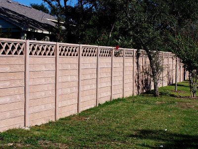 A long, light brown wooden privacy fence with a decorative lattice top runs alongside a grassy yard with several small trees and a house partially visible in the background.