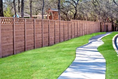 A tall wooden privacy fence with a lattice top runs alongside a winding concrete sidewalk bordered by neatly trimmed green grass and trees in the background.